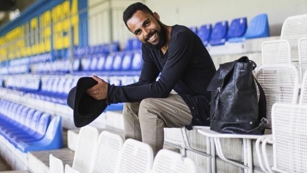 Ein lächelnder Mann mit Hut sitzt auf der Tribüne eines Stadions.