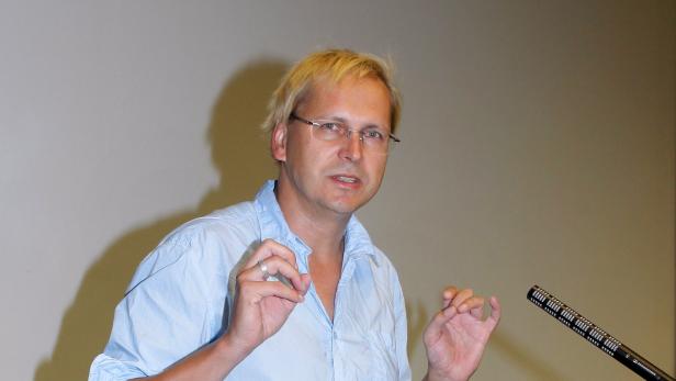 Ein Mann mit blonden Haaren und Brille gestikuliert vor einem Mikrofon.