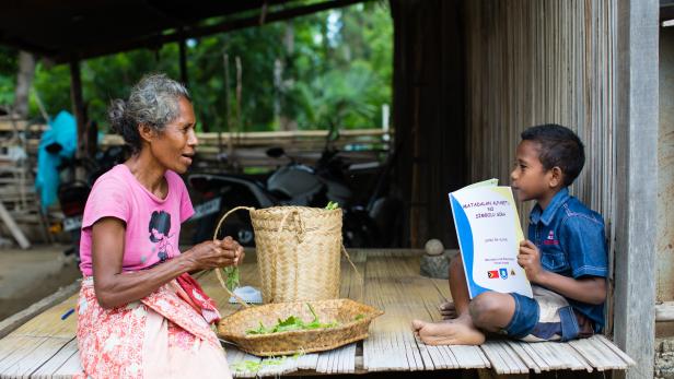 Ein Bub erzählt seiner Großmutter, was in dem Schulbuch steht, aus dem er vor seiner Hütte in Osttimor lernt