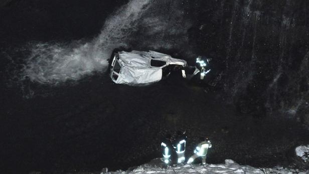 Ein Auto liegt in einem Fluss, während Feuerwehrleute am Ufer stehen.