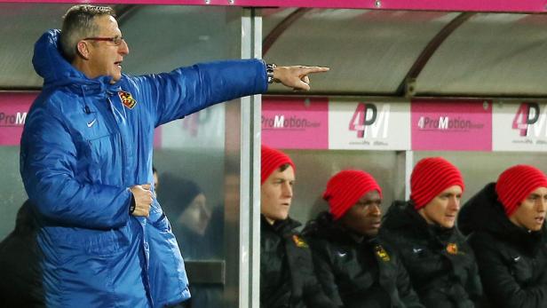 Ein Trainer in blauer Jacke zeigt auf dem Spielfeld, während Spieler mit roten Mützen auf der Bank sitzen.