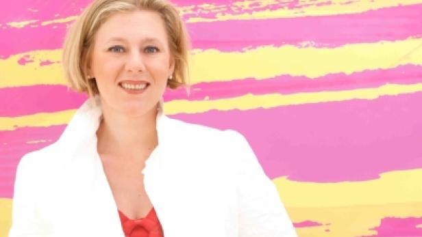 Eine Frau mit blonden Haaren lächelt vor einem farbenfrohen Hintergrund.