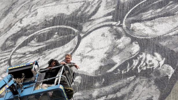 Zwei Personen stehen in einer Hebebühne vor einem großen Wandgemälde.