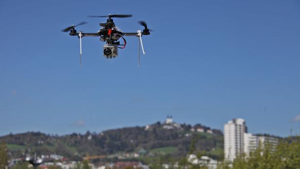 Eine Drohne mit Kamera fliegt vor blauem Himmel über einer Stadt.