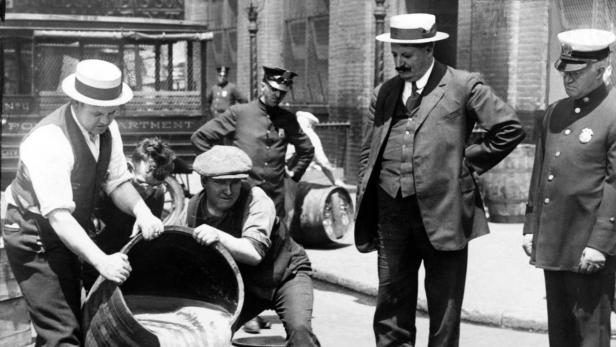 Zwei Männer leeren ein Fass mit Flüssigkeit in einen Gully, während andere Männer zusehen.