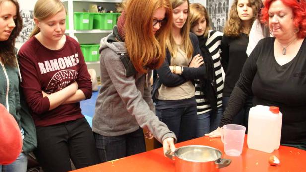 Eine Gruppe von Schülerinnen beobachtet, wie eine rothaarige Schülerin einen Topf auf einen Tisch stellt.