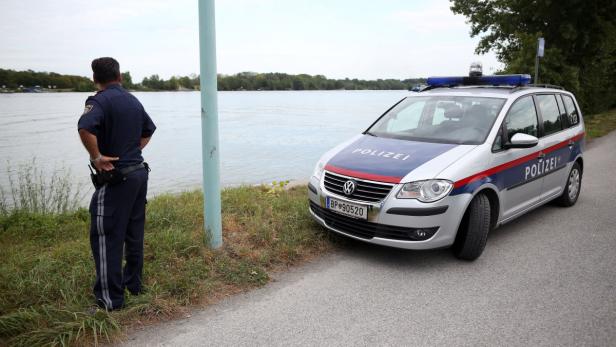 Ein österreichischer Polizist steht neben einem Polizeiauto am Ufer eines Sees.
