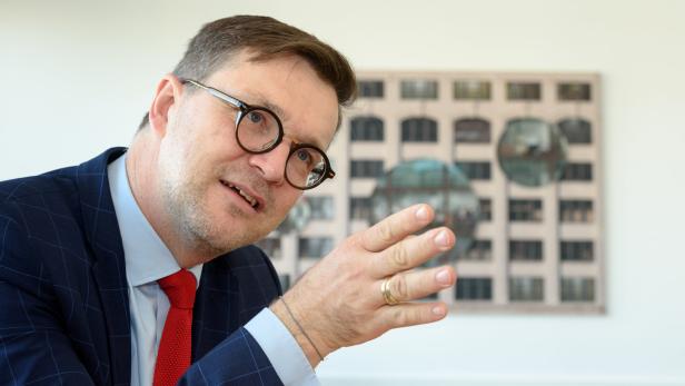 Ein Mann mit Brille, Anzug und roter Krawatte gestikuliert vor einem Bild.