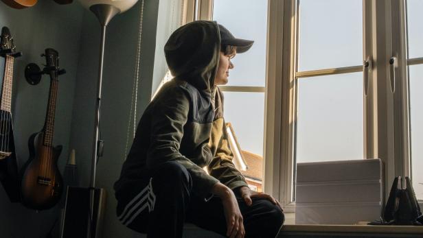 Ein Jugendlicher mit Kapuzenpullover sitzt auf einer Fensterbank und schaut nach draußen.