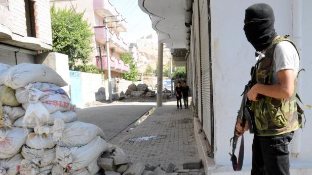 Ein maskierter Mann mit Gewehr steht auf einer Straße, die mit Sandsäcken und Trümmern blockiert ist.