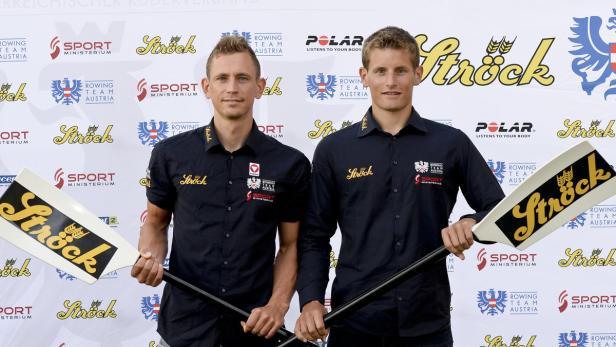 Zwei Ruderer des österreichischen Nationalteams posieren mit ihren Rudern vor einer Sponsorenwand.