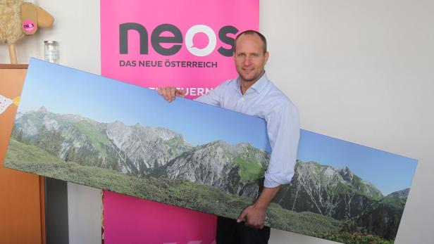 Ein Mann hält ein Panoramabild einer Berglandschaft vor einem NEOS-Hintergrund.