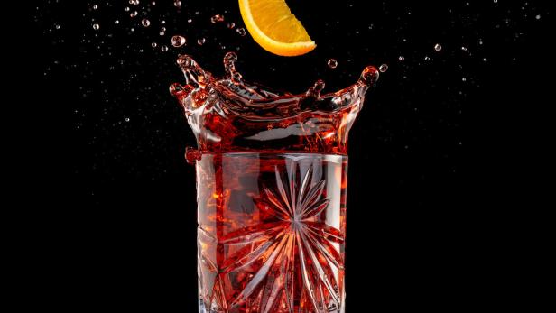 Ein Orangenschnitz fällt in ein Glas mit roter Flüssigkeit und erzeugt eine Fontäne.