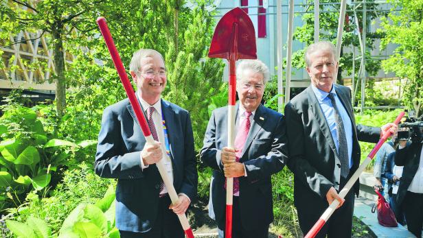 Drei Männer in Anzügen halten Spaten für einen Spatenstich bereit.
