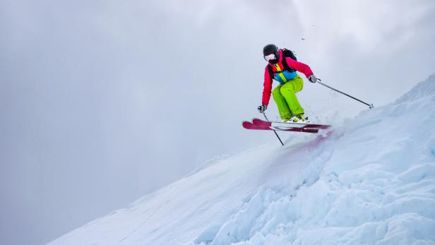 A girl in a green ski suit jumps from a snowy ledge against a cloudy sky in cloudy weather. Sun gleams through the clouds. The concept of winter sports and extreme