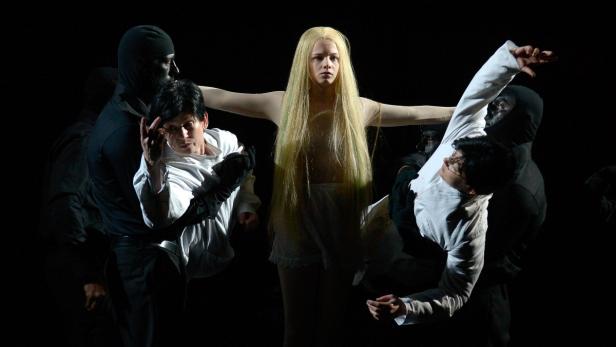 Eine Frau mit blonden Haaren steht mit ausgestreckten Armen, während andere Personen sie zu beiden Seiten stützen.