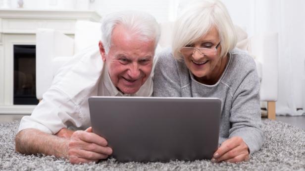 Ein älteres Paar liegt auf einem Teppich und schaut lächelnd auf ein Tablet.
