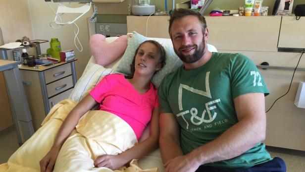 Eine Frau liegt in einem Krankenhausbett, während ein Mann neben ihr sitzt.