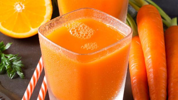 Zwei Gläser frisch gepresster Karottensaft mit Karotten, Orange und Kurkuma auf einem Tablett.