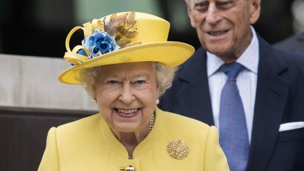 Königin Elisabeth II. lächelt in einem gelben Kostüm mit Blumenstrauß, während Prinz Philip im Hintergrund steht.