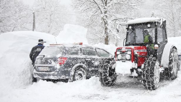 Ein Mann schaufelt Schnee von einem Auto, während ein Traktor mit Schneeketten bereitsteht.