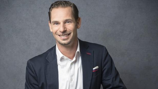 Ein lächelnder Mann in einem dunklen Anzug und einem weißen Hemd vor einem grauen Hintergrund.