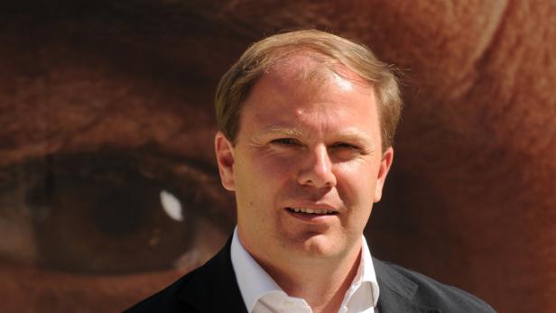 Ein Mann mit blonden Haaren und dunklem Anzug vor dem Hintergrund eines riesigen Auges.