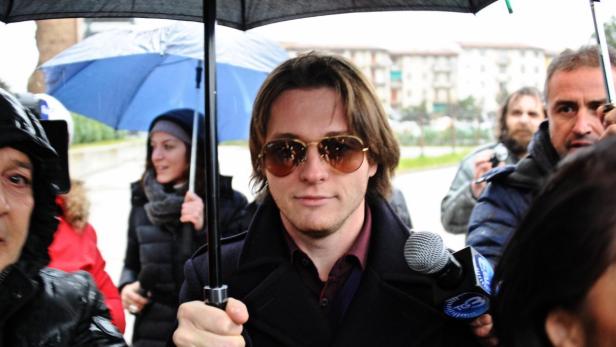Ein Mann mit Sonnenbrille wird von Reportern unter Regenschirmen umringt.