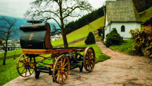 Ein historischer, pferdeloser Wagen steht auf einem gepflasterten Weg.