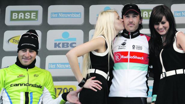 Ein Radrennfahrer wird auf dem Podium von zwei Frauen flankiert und von einer geküsst.