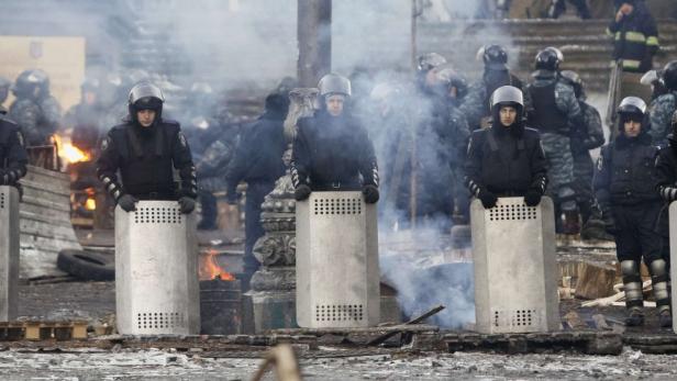 Eine Reihe von Polizisten mit Schutzschilden steht vor Rauch und Trümmern.