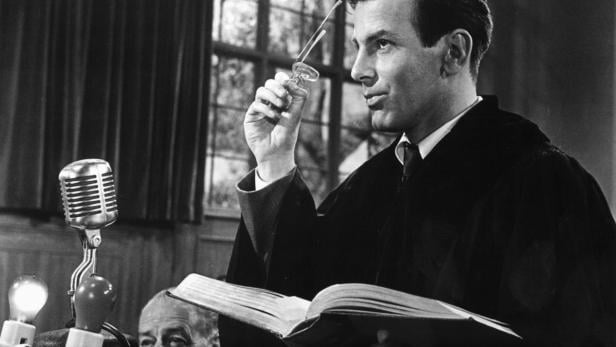 Ein Mann im Talar hält eine Brille in der Hand und liest aus einem Buch.