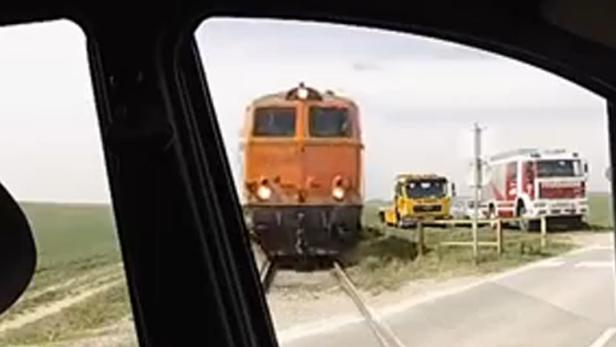 Blick aus einem Auto auf einen Bahnübergang mit einem Zug, einem gelben LKW und einem Feuerwehrauto.