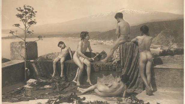 Eine Gruppe nackter junger Menschen posiert vor einer Landschaft mit einem Vulkan im Hintergrund.