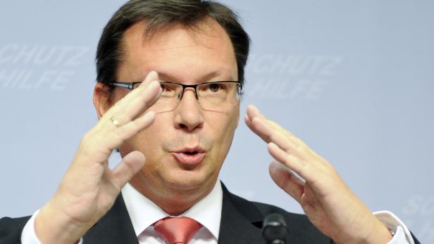 Ein Mann mit Brille gestikuliert vor einem hellblauen Hintergrund.