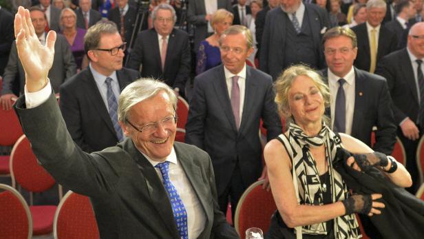 Ein Mann im Anzug hebt lachend die Hand, während eine Frau neben ihm steht.