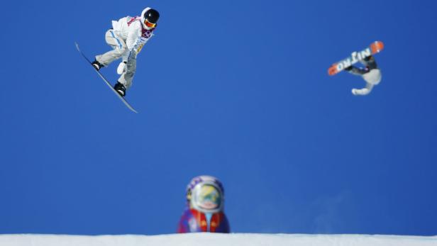 Zwei Snowboarder springen vor blauem Himmel, im Vordergrund eine Matroschka-Puppe.