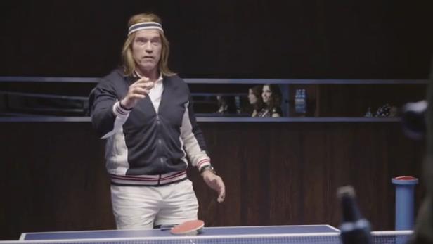 Arnold Schwarzenegger in einer Perücke spielt Tischtennis.