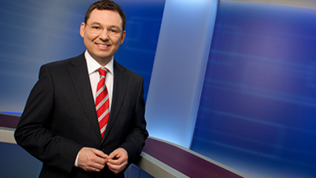 Ein Mann mit Brille, Anzug und Krawatte lächelt vor einem blauen Hintergrund.