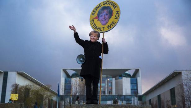 Eine Frau mit Merkel-Maske hält ein Schild mit der Aufschrift „Keine Sonne strahlt wie ich“.