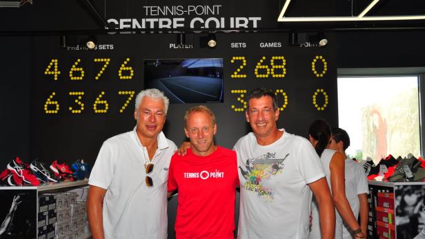 Drei Männer posieren vor einer Anzeigetafel in einem Tennis-Point-Geschäft.