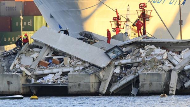 Einsatzkräfte untersuchen die Trümmer eines eingestürzten Gebäudes am Hafen, im Hintergrund ein Schiff.