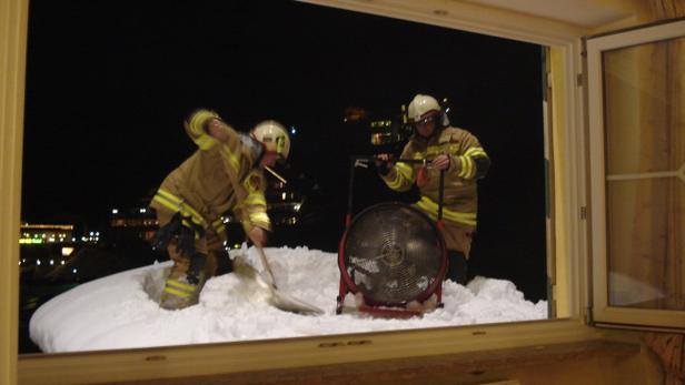Zwei Feuerwehrleute schaufeln Schnee von einem Dach, während ein Ventilator bereitsteht.