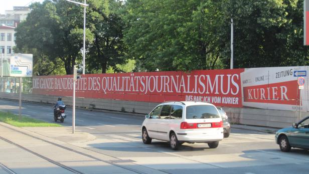 Ein weißer Van fährt auf einer Straße in Wien vor einer Werbung des „Kurier“.