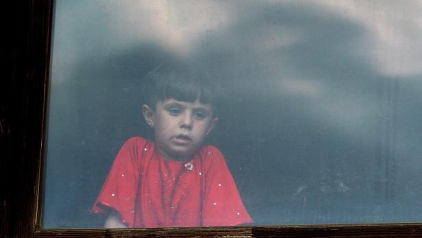 Ein kleines Mädchen mit rotem Oberteil schaut aus einem Fenster.