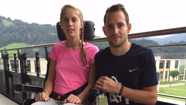 Eine junge Frau im Rollstuhl und ein Mann sitzen auf einem Balkon mit Blick auf eine bewaldete Hügellandschaft.