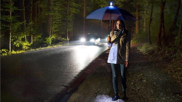 Eine Frau steht nachts mit einem leuchtenden Regenschirm an einer dunklen Straße.