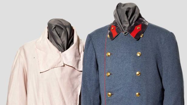 Zwei Uniformjacken, eine hellrosa und eine blaue mit goldenen Knöpfen, stehen nebeneinander.