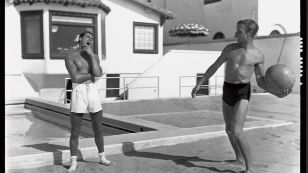 Zwei Männer in Badehosen posieren an einem Pool.