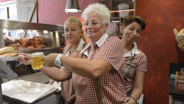 Drei Frauen in Trachtenhemden arbeiten in einem Imbiss mit Fleisch und Bier.